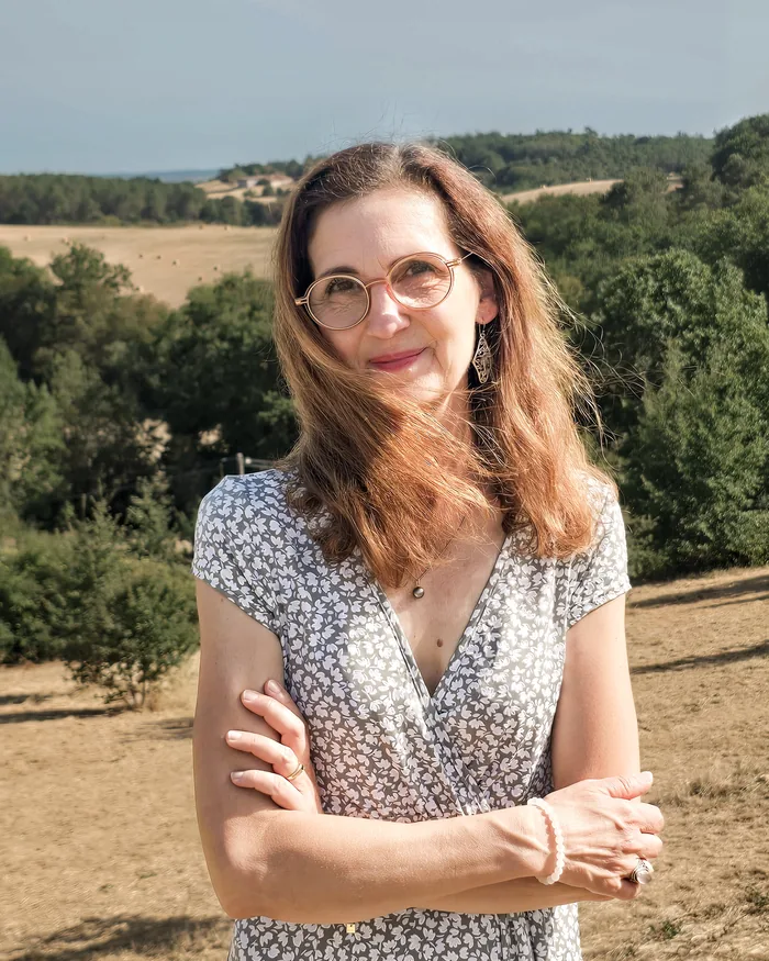 Christine BARROS, artiste peintre, bras croisés et sourire, devant les collines boisées et les champs de la campagne de Dordogne